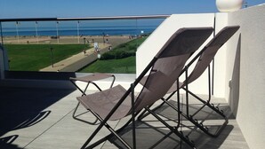 A balcony with two reclining chairs, a white spherical object on the wall, and a view of a beach and promenade.
