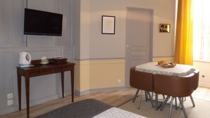 A hotel room with a bed, a TV mounted on the wall, a small table with chairs, and a window with yellow curtains.