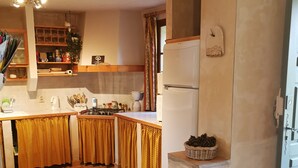 Een keuken met een koelkast, fornuis en diverse keukenbenodigdheden. Op de voorgrond staat een rieten stoel.