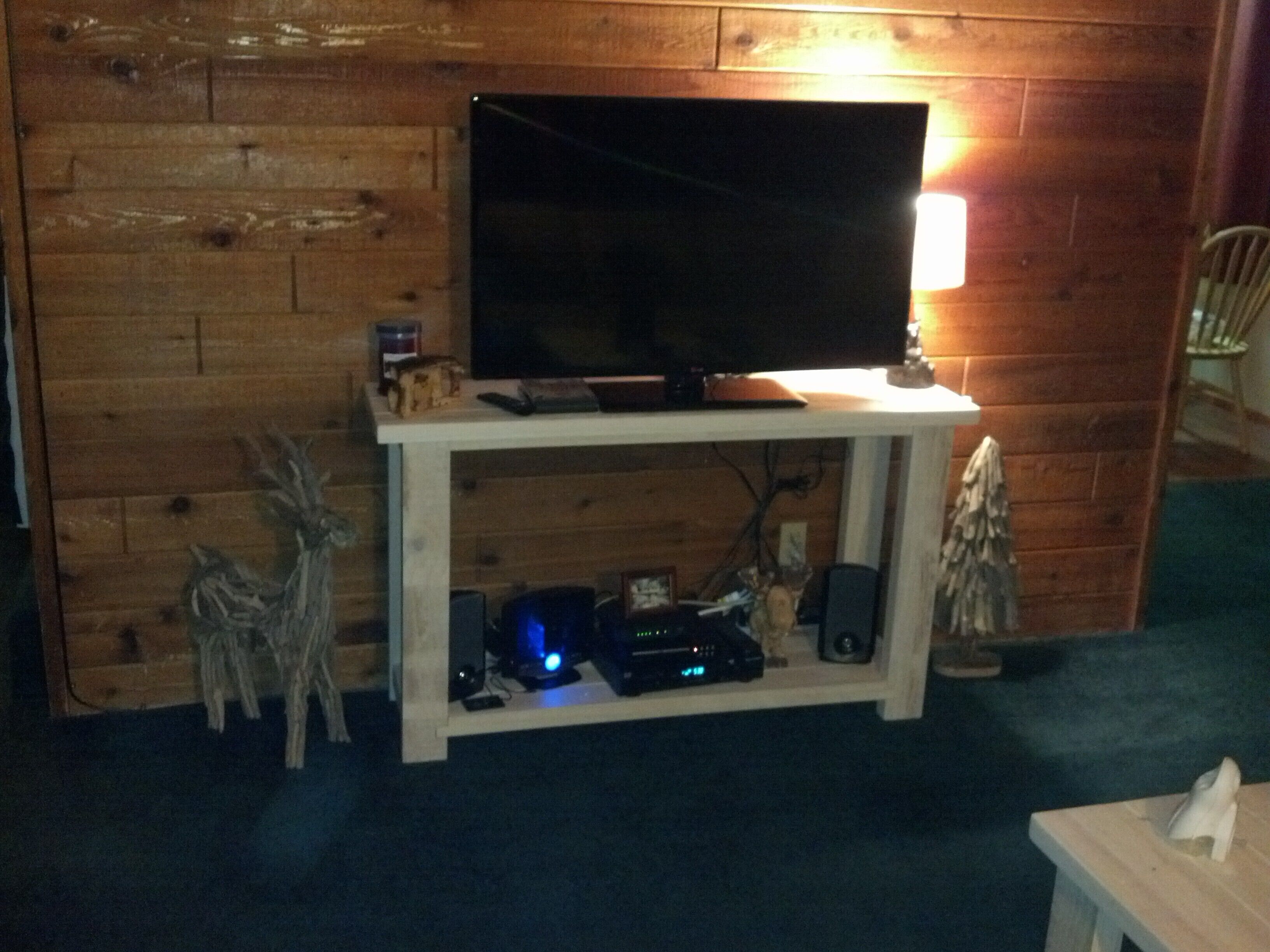 Pared de madera con un televisor sobre un soporte blanco, un estante pequeño con dispositivos electrónicos y una silla al fondo.