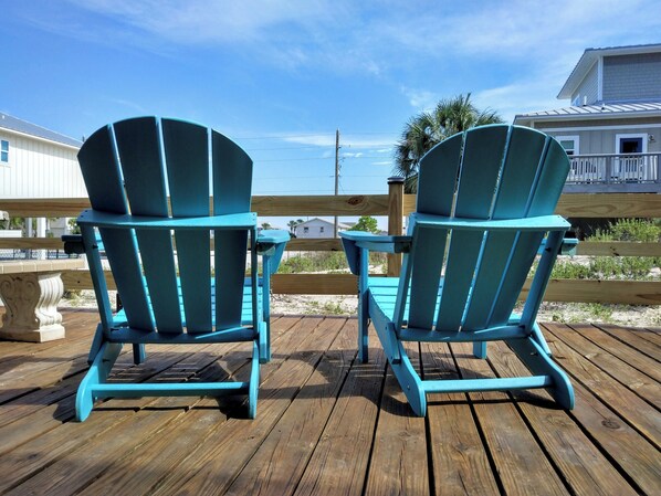 Two turquoise Adirondack chairs on a wooden deck with a view of a beach and houses in the background.