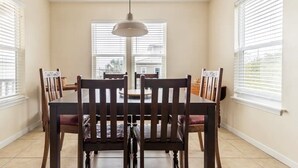 A dining room with a round table and six chairs, large windows with blinds, and a hanging pendant light. - A secluded 3 bedroom, 2 bath Gulf view home nestled in lush tropical vegetation (St George Island)