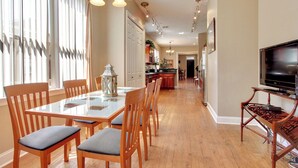 Un grand espace à manger avec une table à plateau en verre et des chaises en bois, une cuisine équipée d’appareils électroménagers modernes et un téléviseur posé sur un meuble en bois.