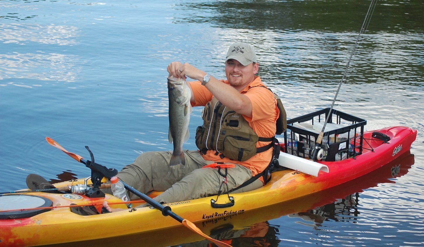 Une personne dans un kayak, tenant un poisson, avec une canne à pêche et une boîte de pêche visibles.