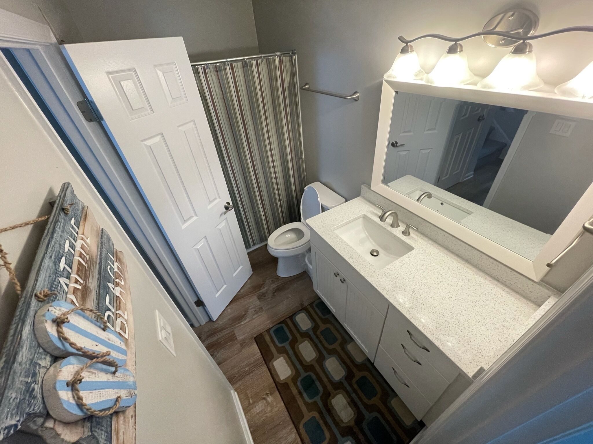 A bathroom with a white sink, a mirror, and a toilet. There is a shower curtain and a patterned rug on the floor.