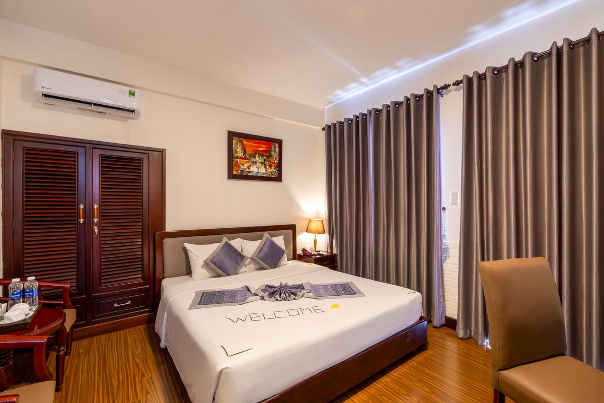 A hotel room with a neatly made bed, a bedside table with a lamp, a chair, and a large window with curtains.