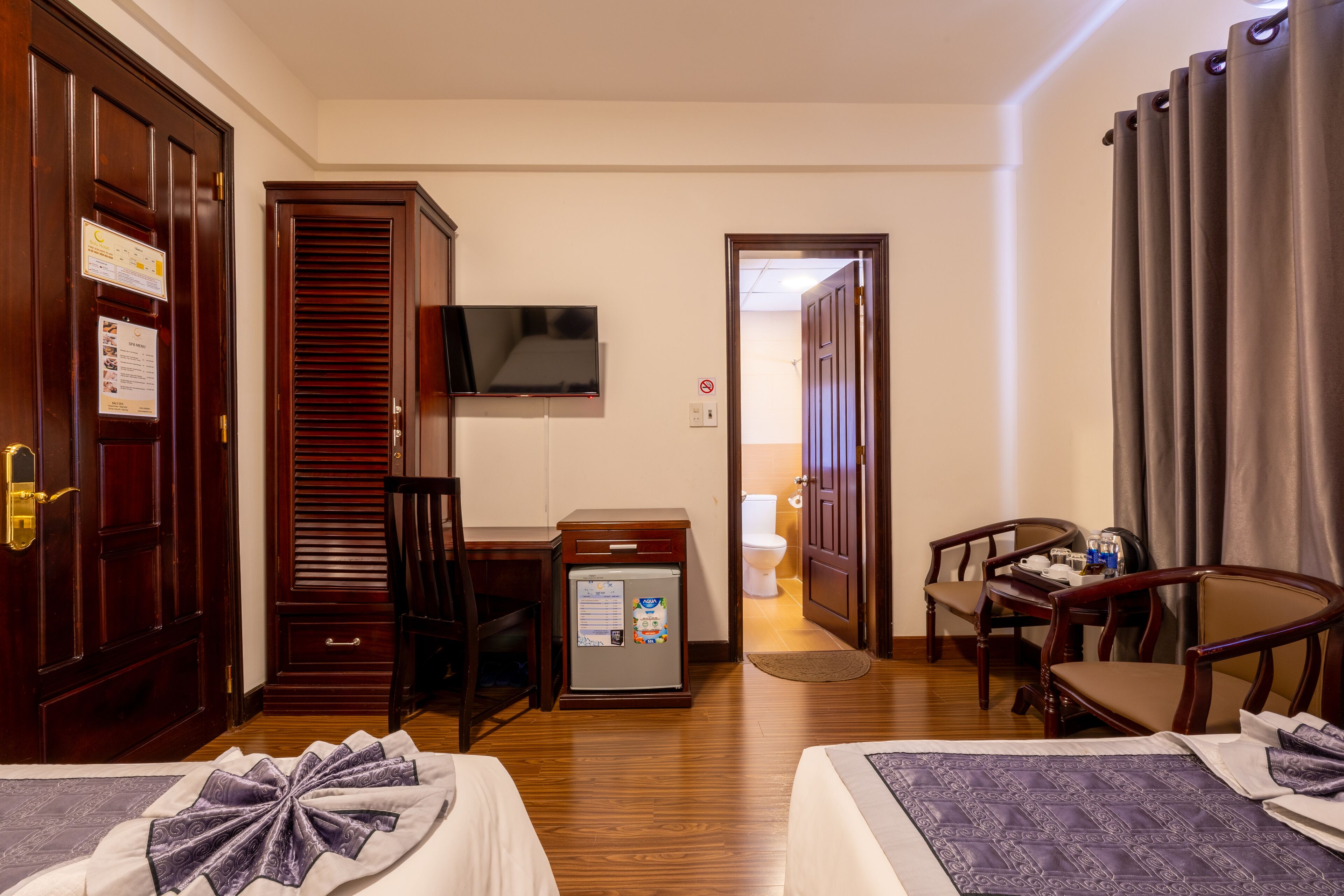 A hotel room with two beds, a desk with a computer, a television, a small refrigerator, and a bathroom visible through an open door.