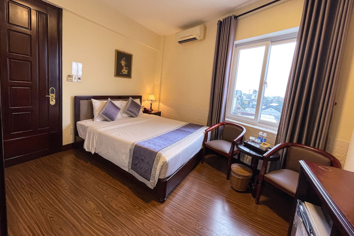 A hotel room with a bed, a desk with a chair, a window with curtains, and a door.