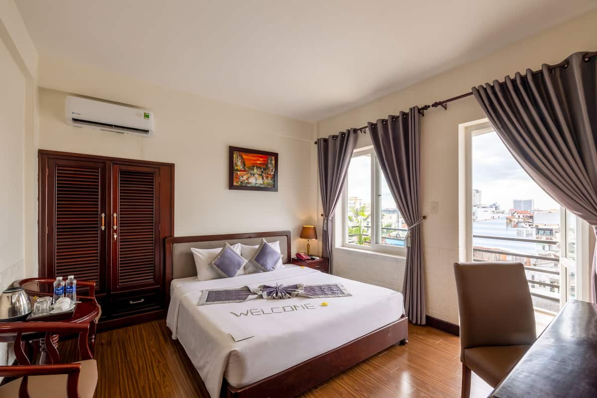 A hotel room with a bed, a desk, a chair, a window with curtains, an air conditioner, and a painting on the wall.