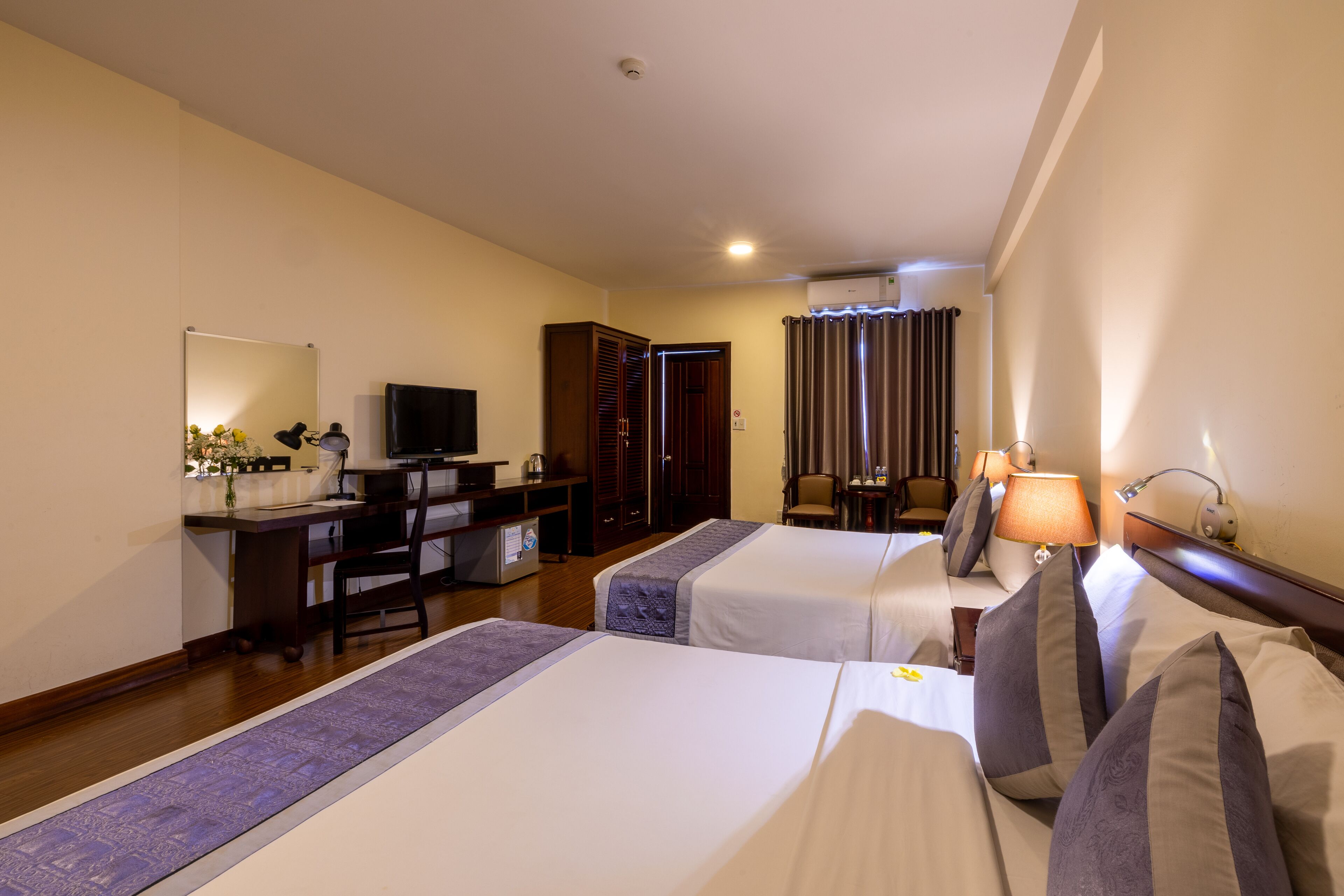A hotel room with two beds, a desk, a television, and an air conditioning unit.