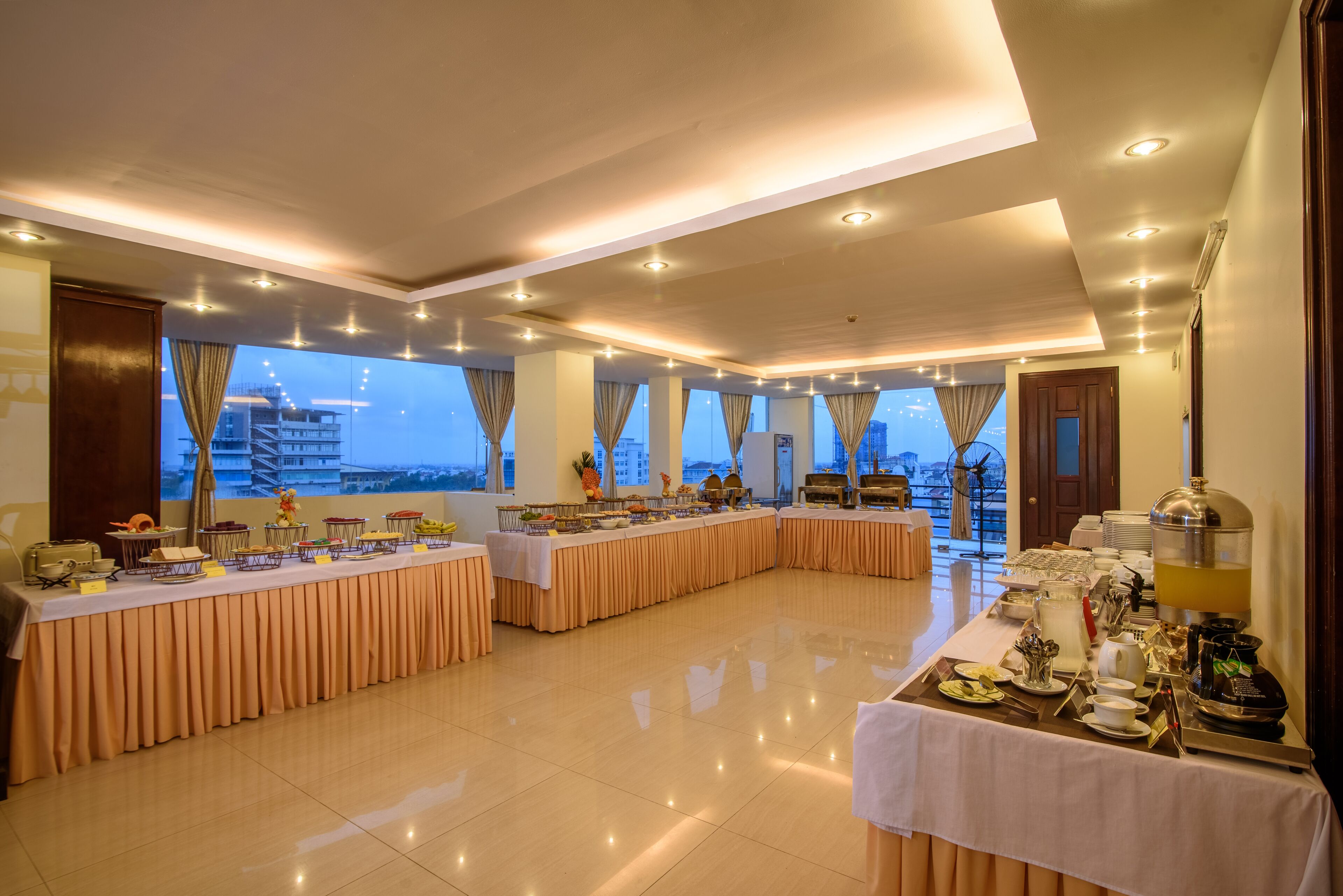 A spacious dining area with multiple tables, a buffet setup, and a view of the cityscape through large windows.