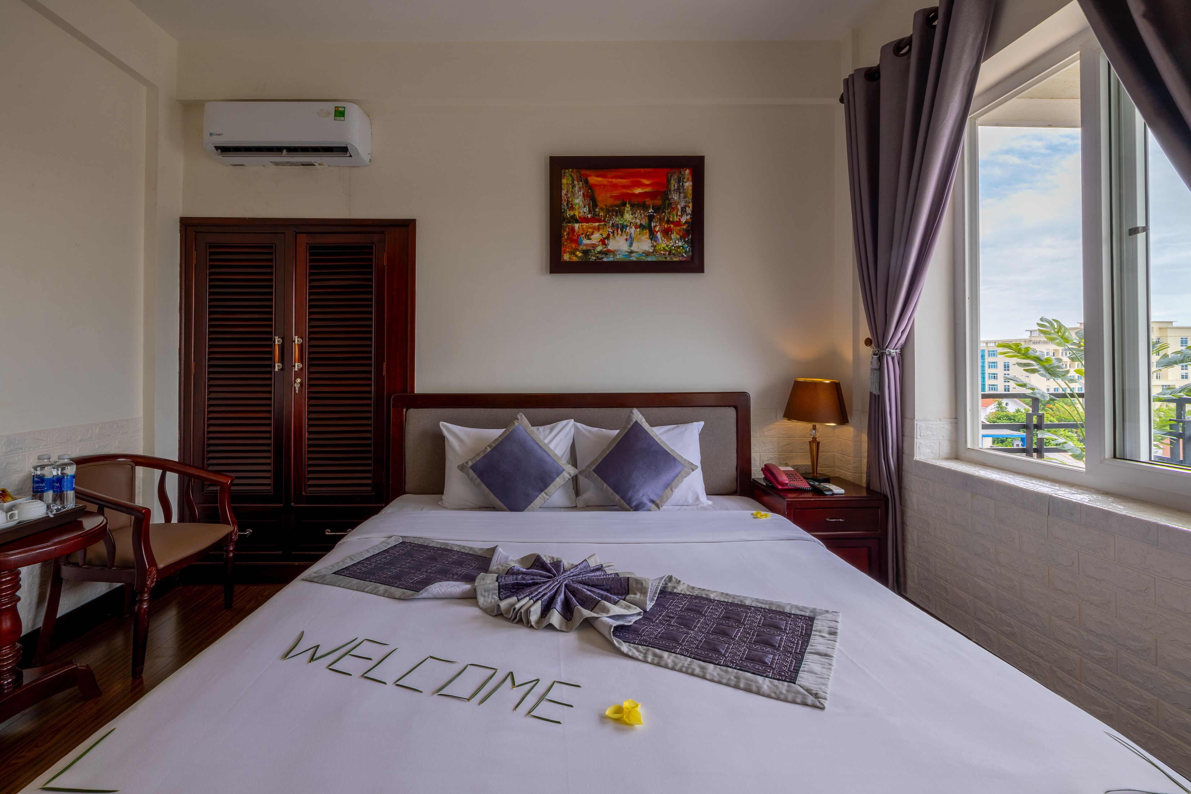 Habitación de hotel con cama, escritorio, silla, mesita de noche, lámpara, ventana con cortinas, un cuadro en la pared y un equipo de aire acondicionado.