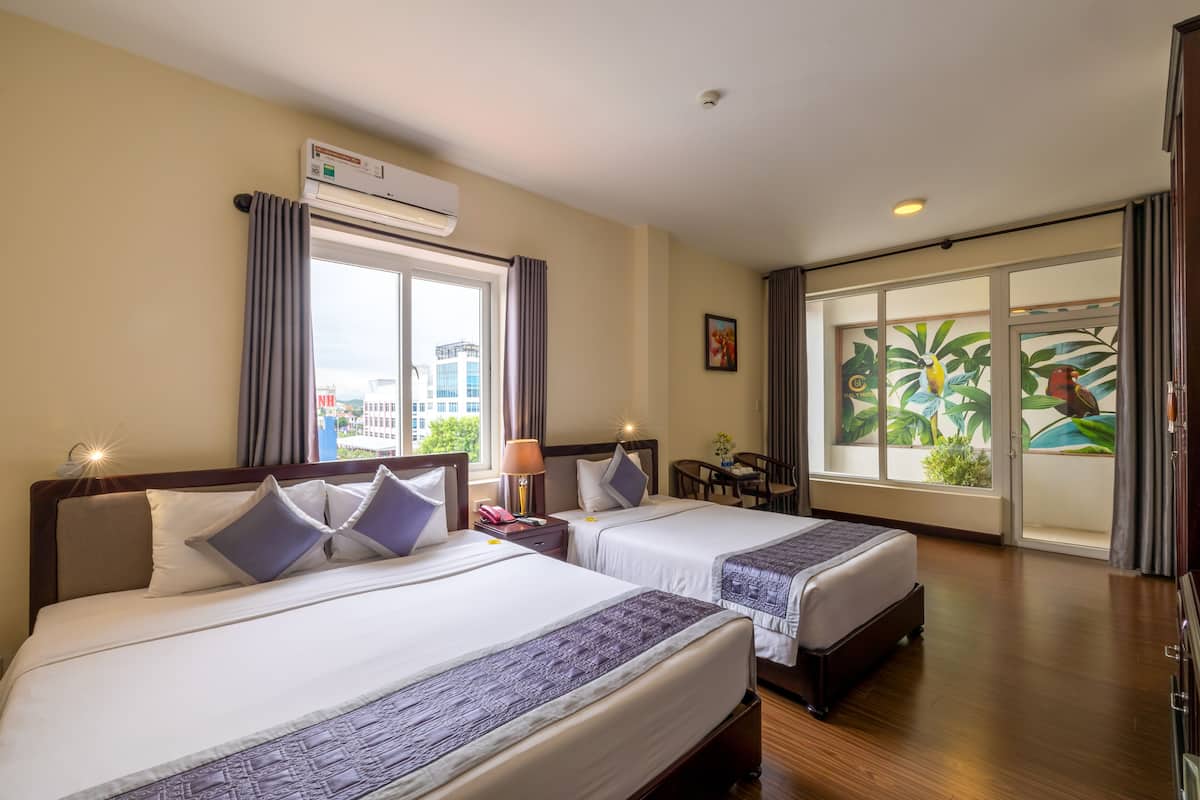 A hotel room with two beds, a desk, a chair, an air conditioning unit, and a large window with a view of buildings and greenery.