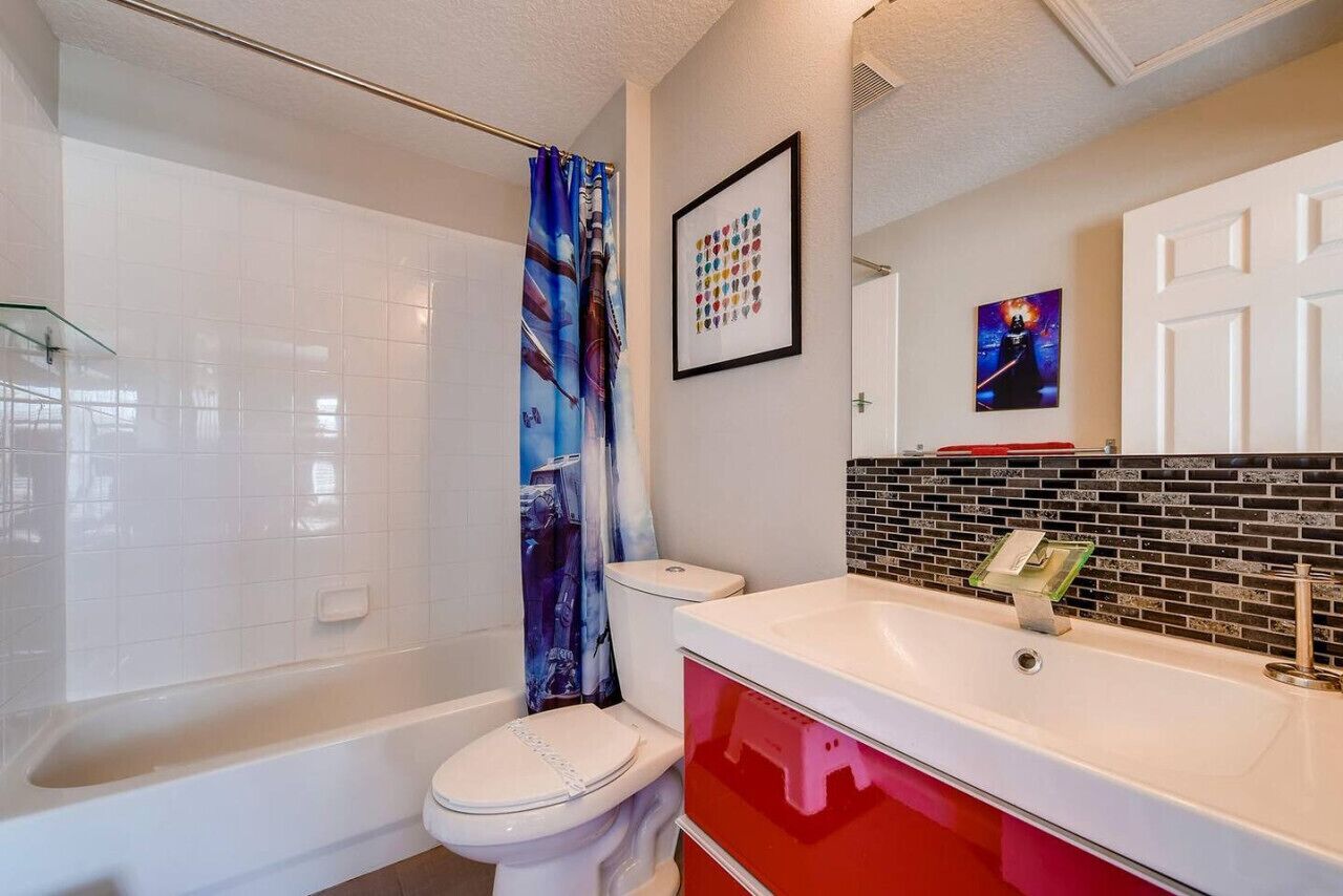 Un baño con inodoro blanco, bañera, cortina de ducha, lavamanos con espejo y pared con azulejos.