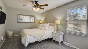 A bedroom with a bed, bedside lamps, a ceiling fan, a window with blinds, a mirror, and a trash can. - ♥Cozy Townhome w Lux Beds♥Full Kitchen♥ Deck♥Park access two doors down♥ (Overland Park)
