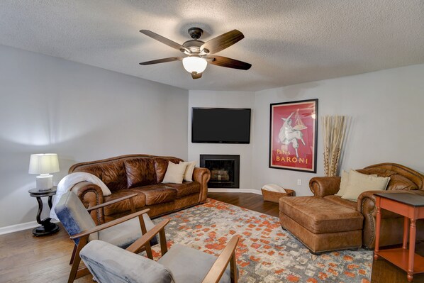 A living room with a brown leather sofa, a wooden armchair, a brown ottoman, a small table, a floor lamp, a fireplace, a ceiling fan, and a mounted TV. - ♥Cozy Townhome w Lux Beds♥Full Kitchen♥ Deck♥Park access two doors down♥ (Overland Park)