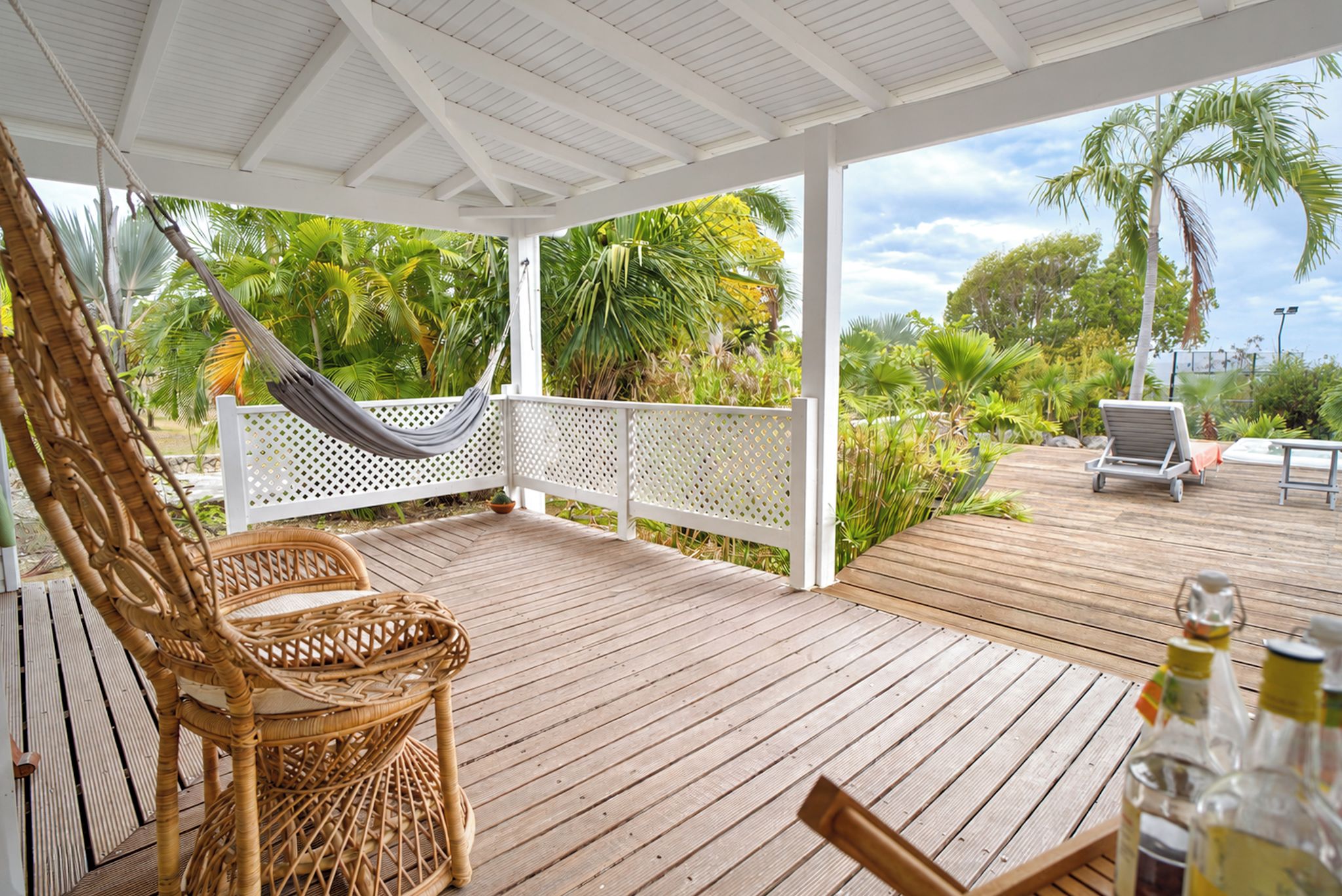 Une terrasse en bois avec une pergola blanche, une chaise en osier, un hamac et une table avec des bouteilles.