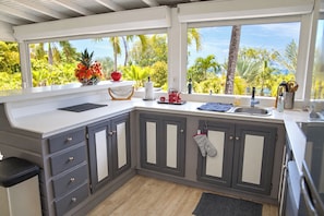 Una cocina con gabinetes grises, encimera blanca y vista a palmeras.