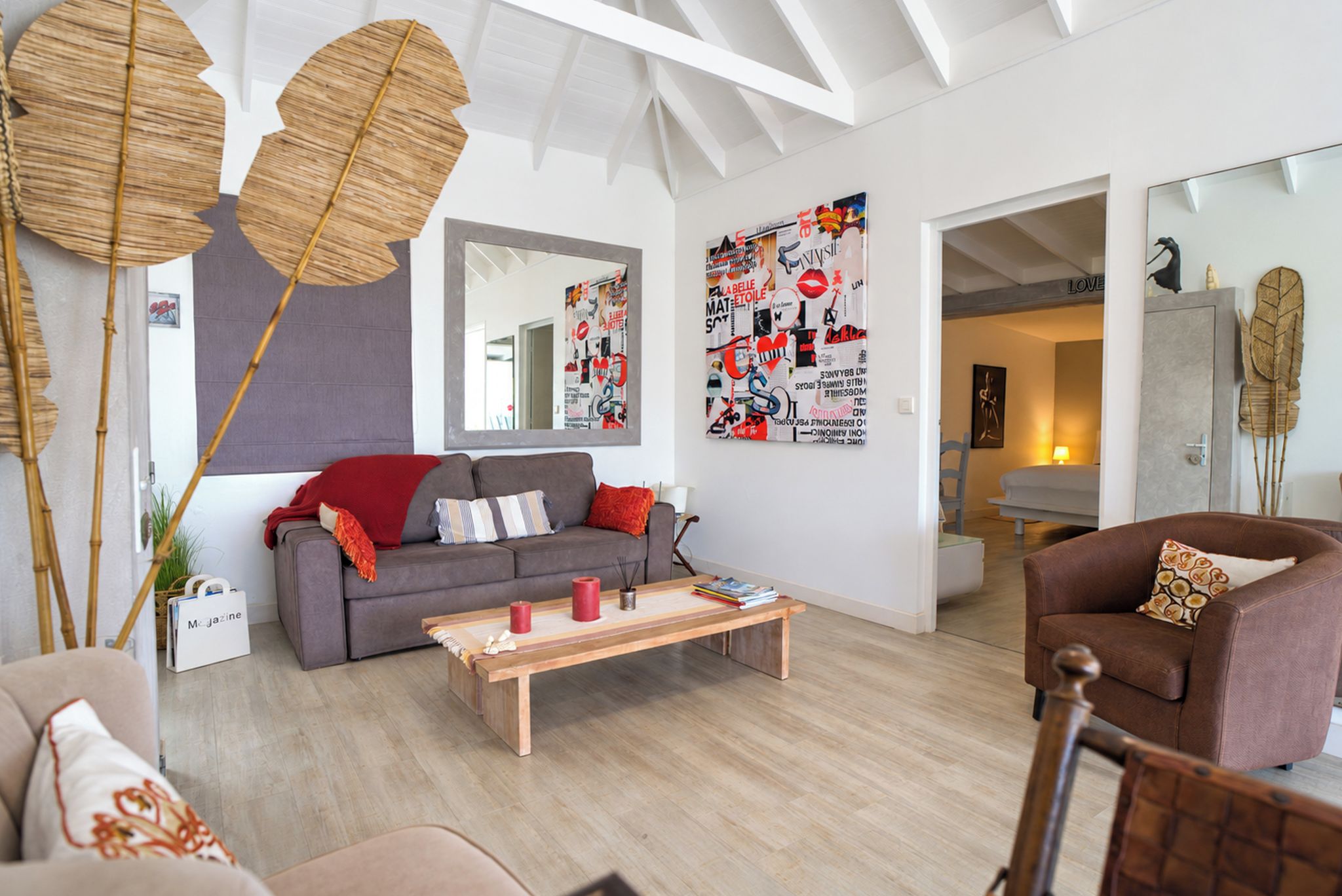 Uma sala de estar moderna com um sofá cinzento, uma mesa de centro de madeira e uma poltrona marrom. Há painéis decorativos nas paredes e um espelho grande.