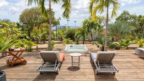 A wooden deck with lounge chairs and a hot tub, surrounded by tropical plants and a view of the sea. - Guest-House In A Palmgrove For A Couple. Space, Privacy And Silence (Saint Martin)