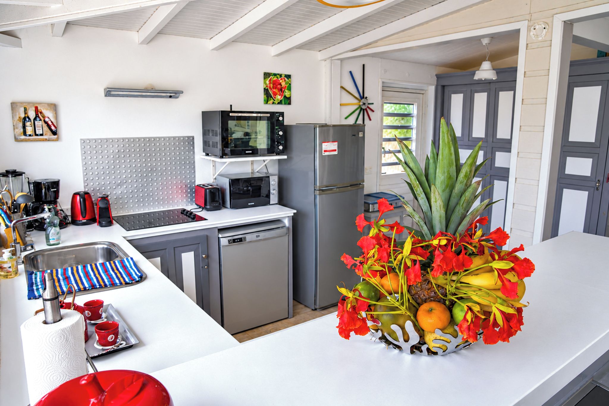 A kitchen with a stainless steel refrigerator, a microwave, and a toaster. There is a sink with a faucet and a dish rack. A vase with red flowers and fruits is on the counter.