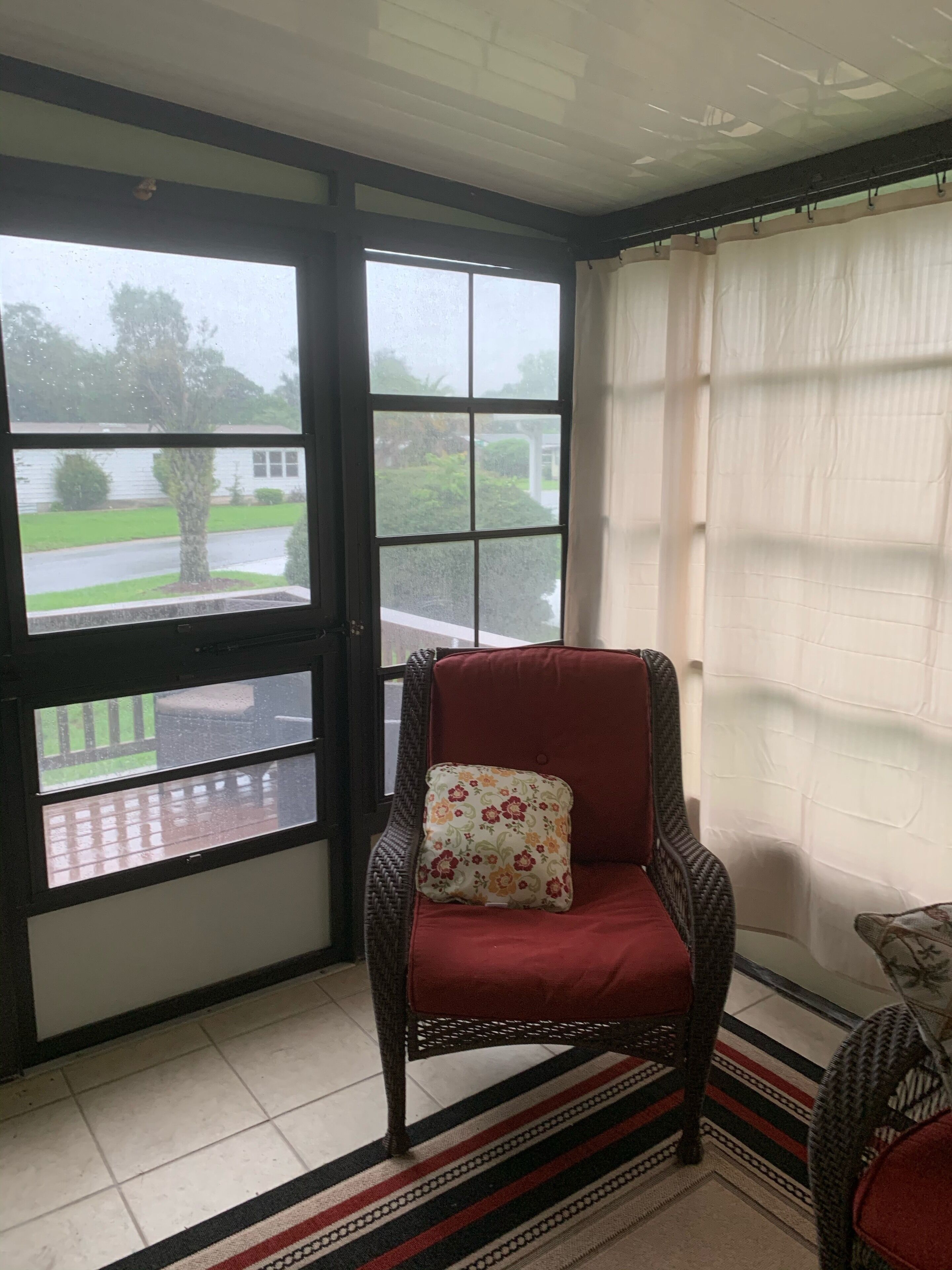 A room with a wicker chair with a floral pillow, a red cushion, and a view of a house and greenery through a large window.