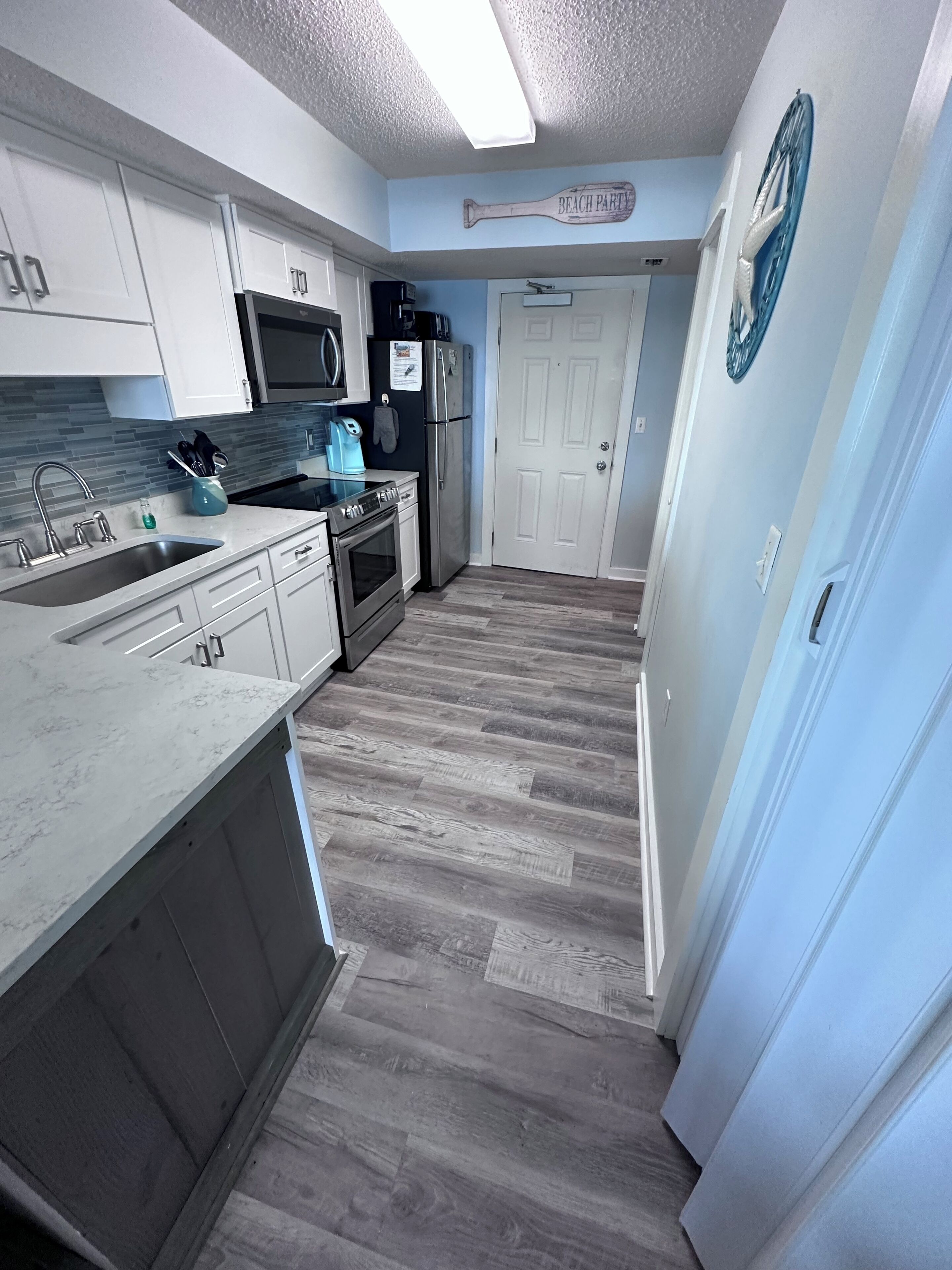 A kitchen with white cabinets, a stainless steel sink, a microwave, a refrigerator, and a stove. There is a wooden floor and a door with a clock on the wall.
