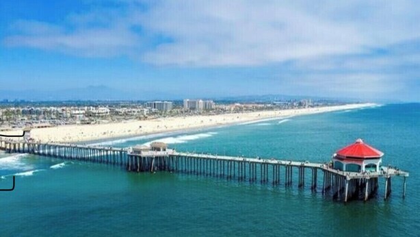 A coastal scene with a pier extending into the ocean, a beach, and a cityscape in the background. - Surf City/Sunset Beach Condo (Huntington Beach)