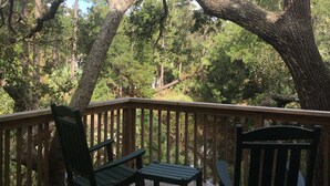 A wooden deck with two rocking chairs and a table, surrounded by large trees and a clear sky.