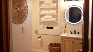 A bathroom with a round mirror, a shelf with folded towels, a wicker basket, and a toilet paper holder.
