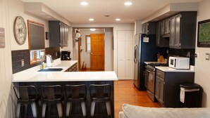 A modern kitchen with a central island, bar stools, dark cabinets, and a microwave.