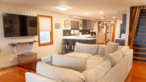 A modern living room with a large beige sectional sofa, a flat-screen TV mounted on the wall, and a kitchen area in the background.