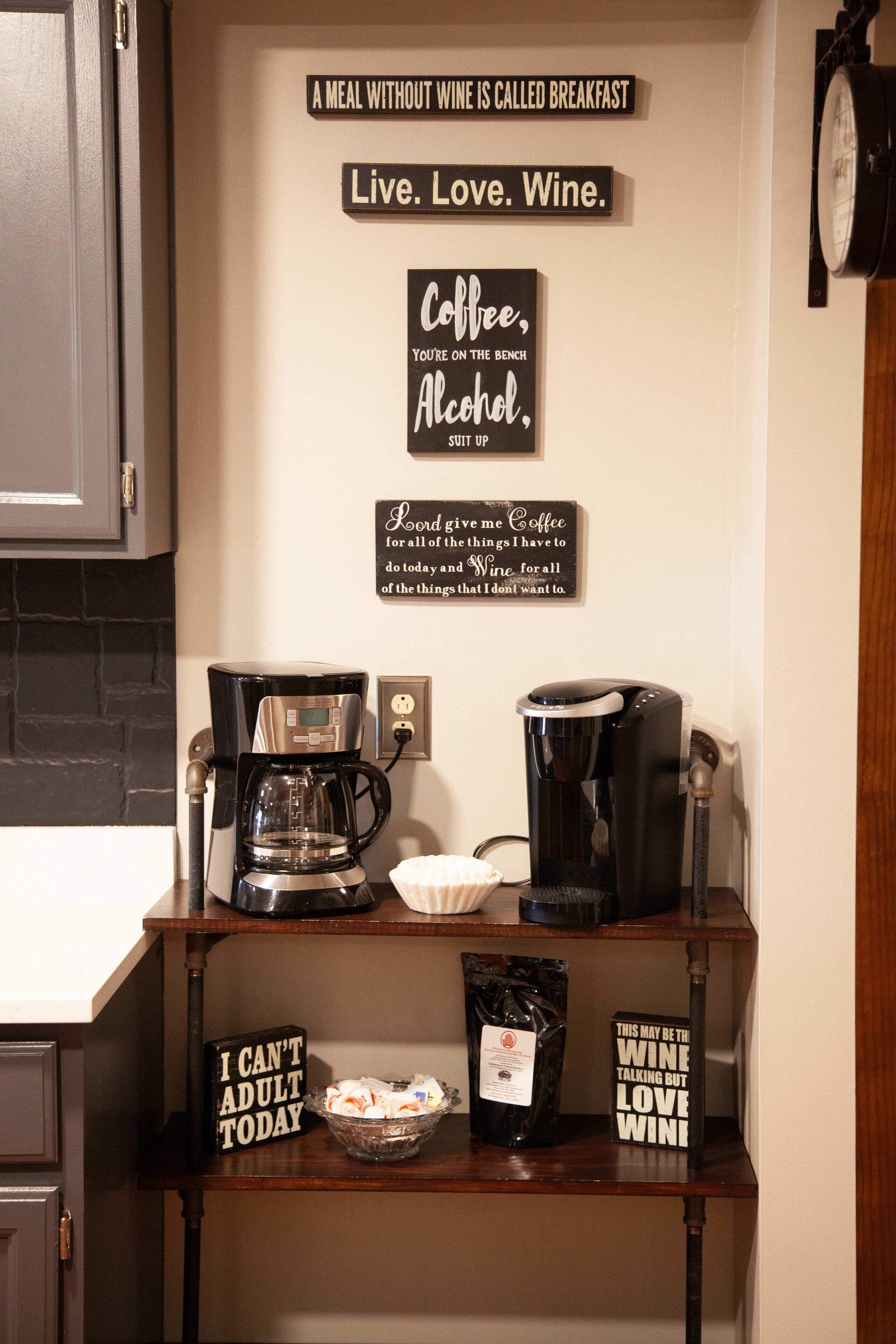 A kitchen counter with coffee makers, a bowl of snacks, and motivational signs about coffee, alcohol, and wine.