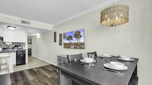 Una cocina moderna con armarios blancos, una mesa de comedor de madera oscura preparada para cuatro personas, y un cuadro grande con palmeras en la pared.