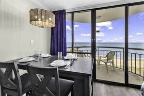 A dining area with a table set for four, a wicker chandelier, and a view of the beach through a glass door.