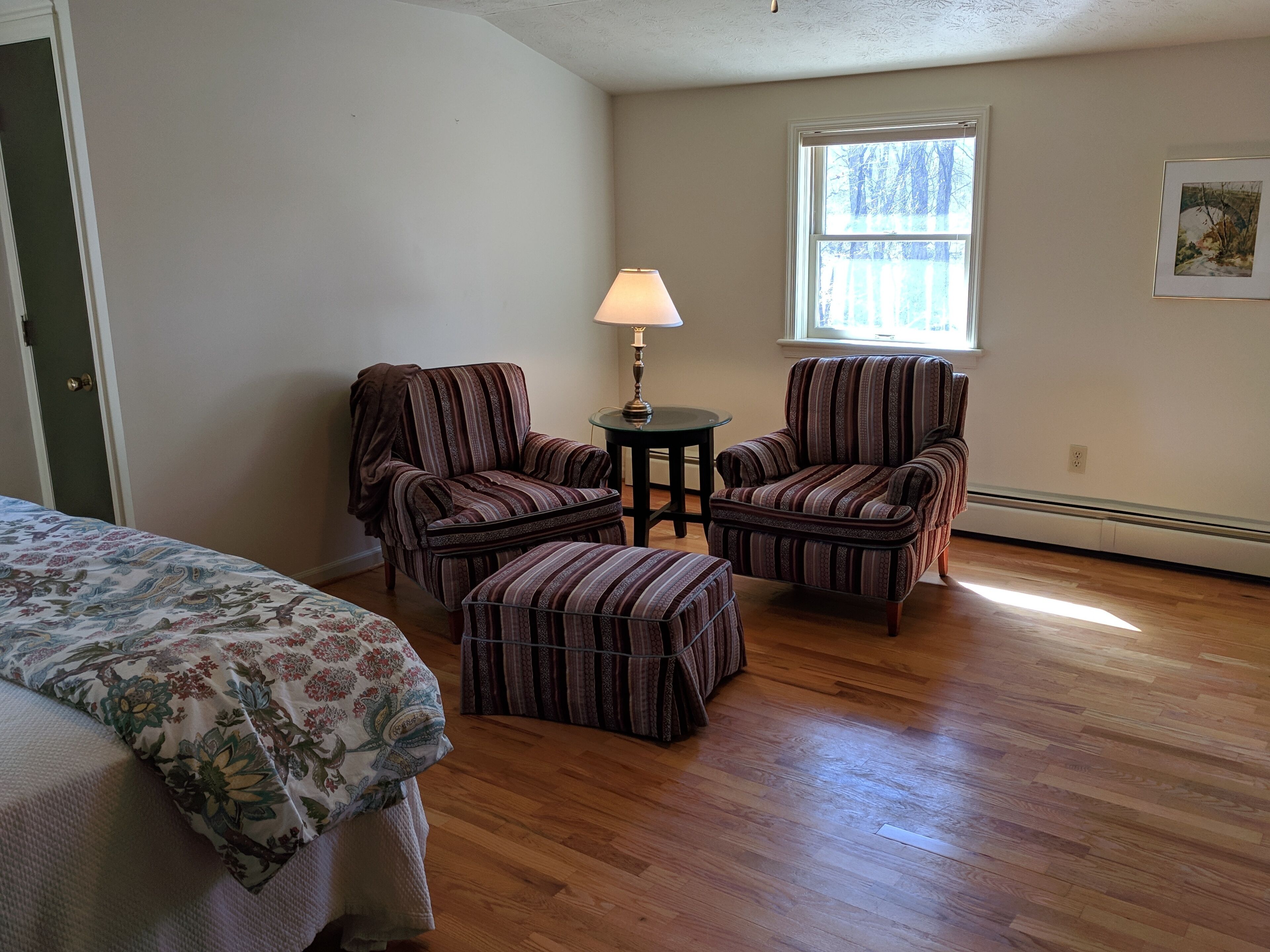Um quarto com piso de madeira, duas poltronas listradas, uma mesa pequena com abajur e uma cama com roupa de cama floral.