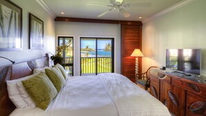 Habitación de hotel con cama, televisor sobre un tocador de madera, ventilador de techo y balcón con vista al océano.