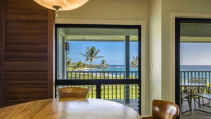 Una mesa y sillas de madera en una habitación con un ventanal que da a un balcón con vistas al océano y palmeras.