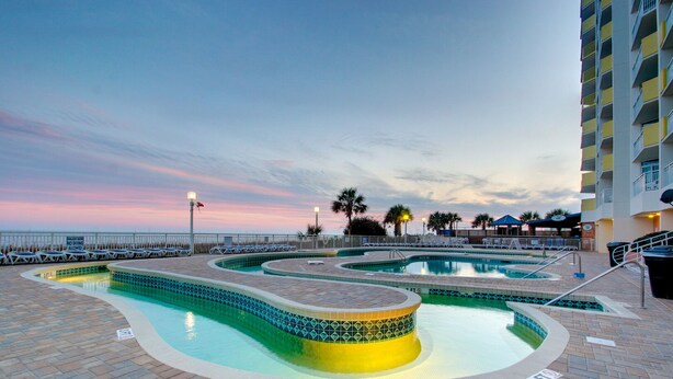 Un área de piscina con varias piscinas, una zona de estar hundida y vistas al océano al atardecer.