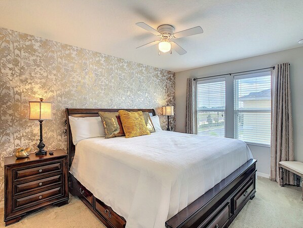 A bedroom with a bed, two bedside lamps, a nightstand, a dresser, a ceiling fan, and a window with curtains.