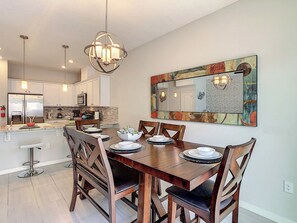 A dining area with a wooden table, chairs, and a chandelier. There is a kitchen in the background with modern appliances.