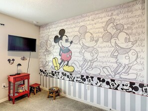 A room with a wall mural featuring Mickey Mouse, a television mounted on the wall, a red table with a toy box, and a small stool.