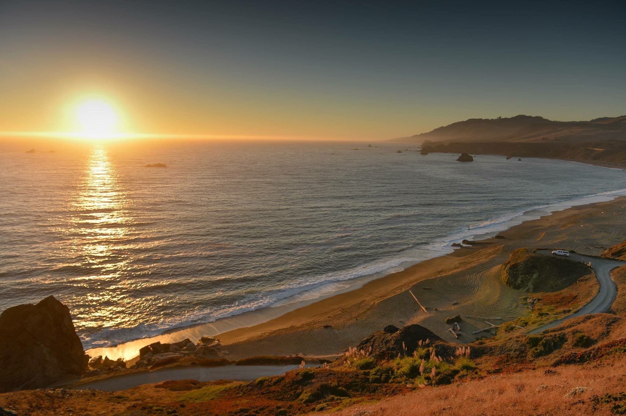 Paisagem costeira com uma estrada sinuosa, afloramentos rochosos e um pôr do sol sobre o oceano.