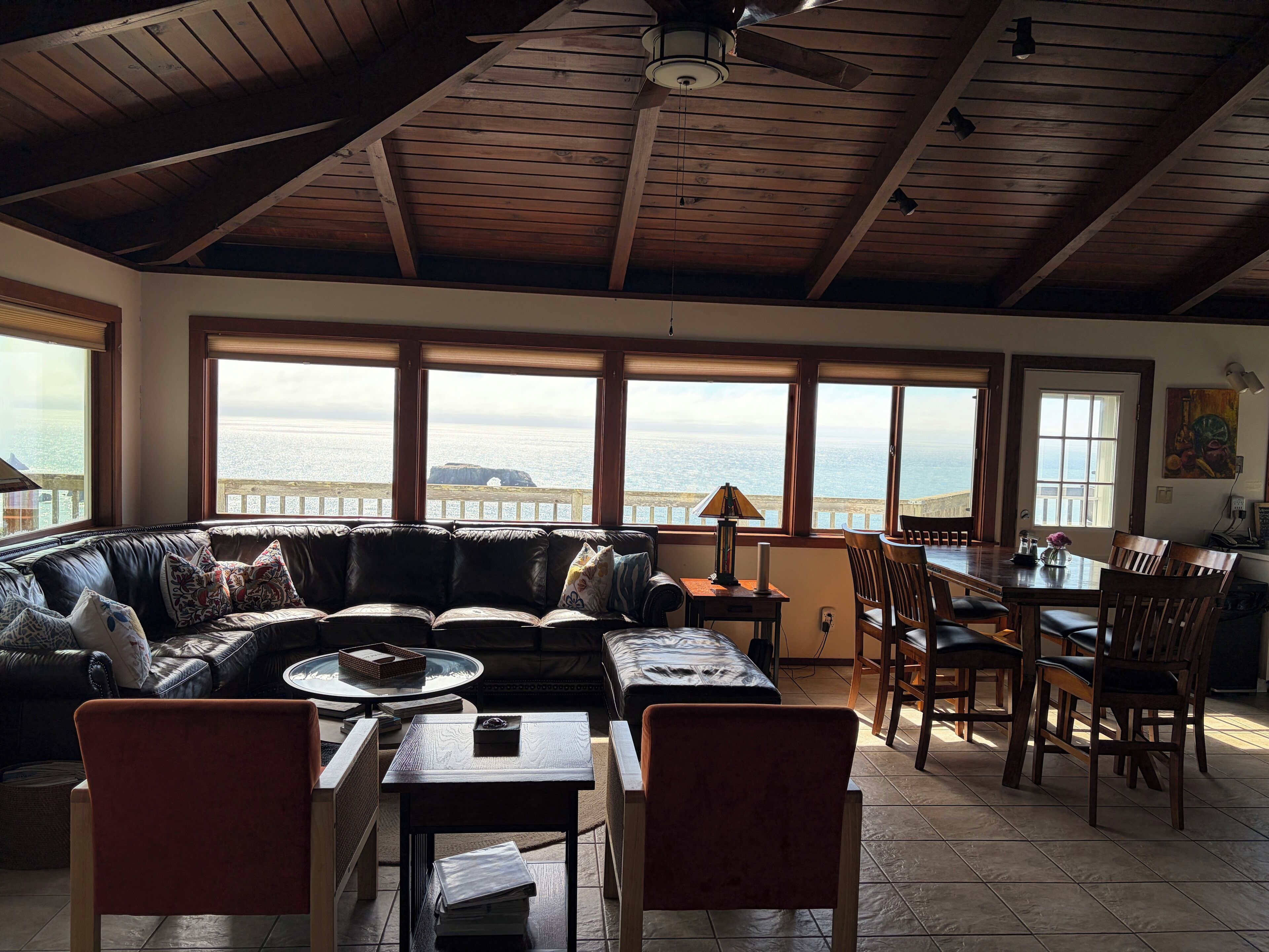 Un salon chaleureux avec une grande fenêtre donnant sur la mer, un plafond en bois et un coin repas avec des chaises et une table.