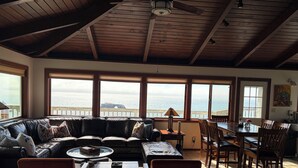Un salon chaleureux avec une grande fenêtre donnant sur la mer, un plafond en bois et un coin repas avec des chaises et une table.