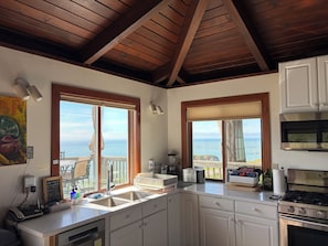 A kitchen with wooden beams, a large window with a view of the sea, a stove, and a sink.