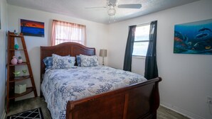 A bedroom with a wooden bed, a ceiling fan, a window with curtains, a painting of a sunset, a painting of an underwater scene, and a wooden ladder with decorative items. - Hippohead Casita Pets stay free bring your Humans! (Homosassa)