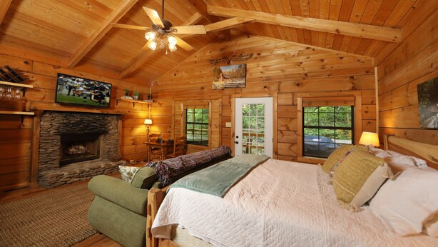 Interior de una cabaña de troncos con chimenea de piedra, una cama con muchos almohadones, un sofá verde y un ventilador de techo.