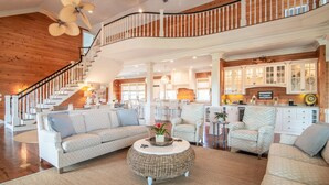 A spacious living area with a high ceiling, wooden beams, a staircase, and a kitchen with white cabinets. - 24 Dene Court Beautiful  8 Bedroom Home in DeBordieu, Private Pool! (Georgetown)