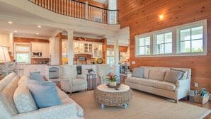 A spacious living area with a high ceiling, wooden paneling, a balcony, and a kitchen in the background. - 24 Dene Court Beautiful  8 Bedroom Home in DeBordieu, Private Pool! (Georgetown)